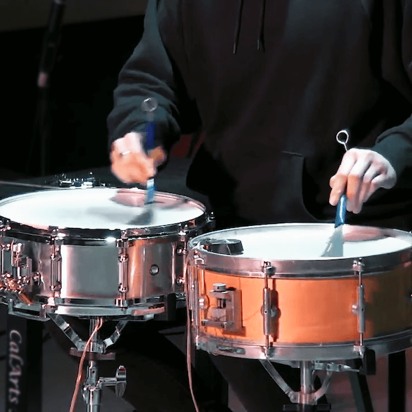 percussionist in black jacket playing two snare drums with blue handled brushes