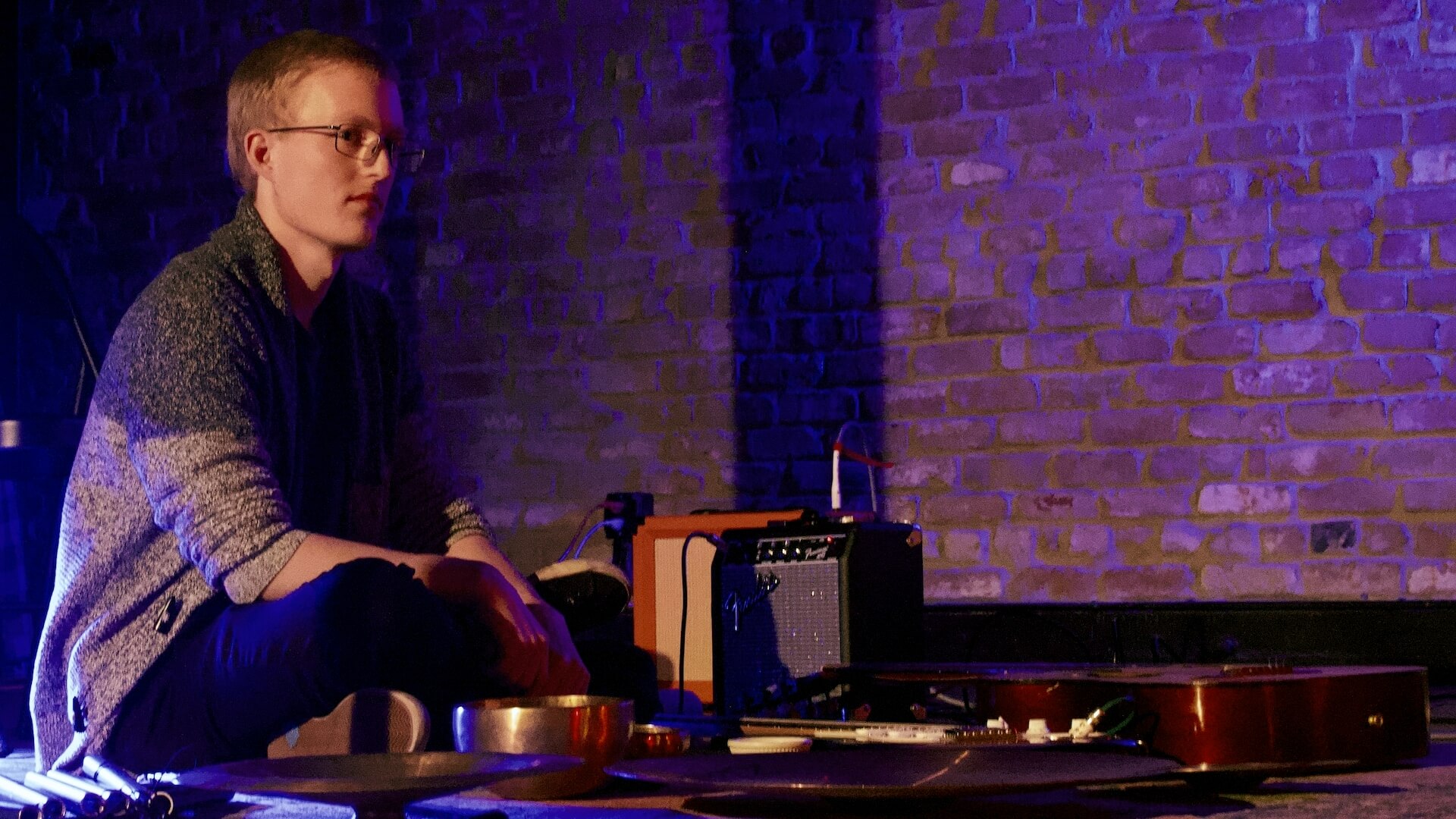 Eric Lennartson sits in front of a wide array of instruments including: singing bowls, wind chimes, cymbals, and two guitars. A purple and blue light is cast on a brick wall background.